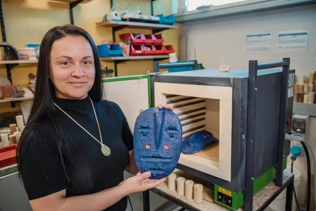 Elena in the ceramics studio holding a blue mask beside the kiln